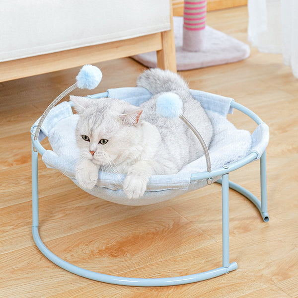 Cat lounging in a small pet bed with pom-poms on a wooden floor.