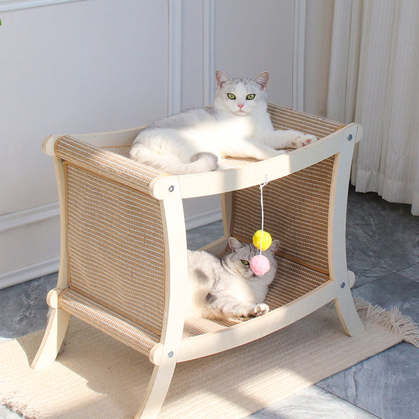Two cats on a wicker pet bed with toys hanging from it, in a room with light-colored walls and a rug.
