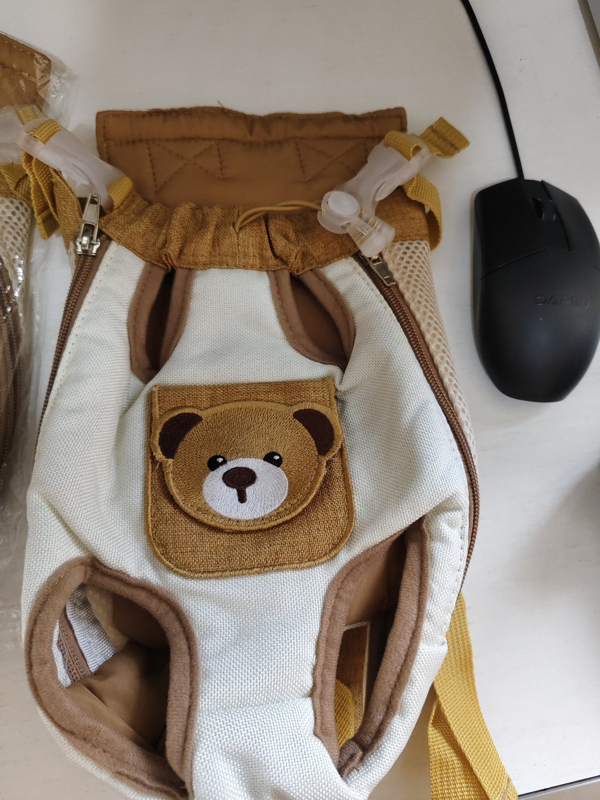 A baby carrier backpack in cream canvas with brown trim and a teddy bear face patch on the front, shown on a light-colored surface next to a black computer mouse.