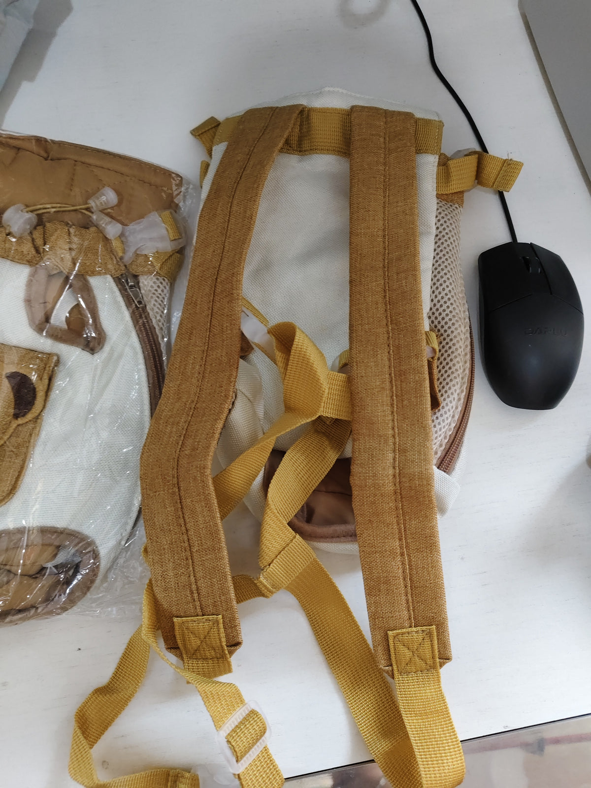 A close-up of a beige and tan backpack shown from the back, featuring adjustable tan webbing straps, mesh side panels, and plastic buckle closures, placed on a white desk next to a black computer mouse.