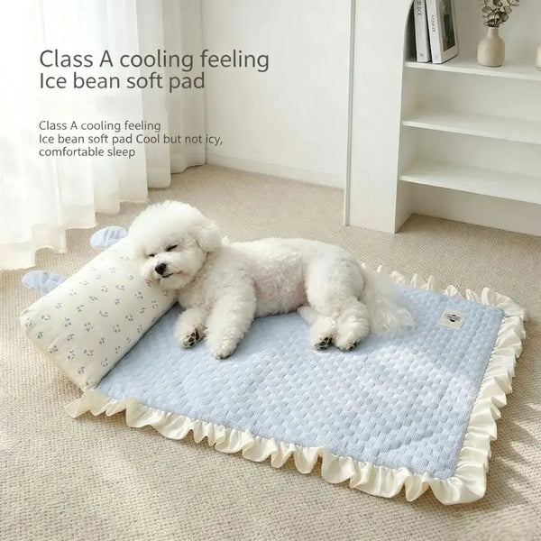 A small white dog lies on a light blue quilted cooling pet pad with cream ruffled edges and a matching pillow in a bright room. Text on the image reads 'Class A cooling feeling Ice bean soft pad'.