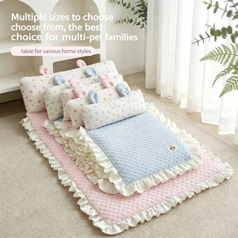 A set of soft baby bedding and pet beds arranged on a beige carpet in a bright room with white curtains and green plants. The set includes a pink ruffled mat, a light blue textured blanket, several white pillows with small prints, and pastel plush toys.
