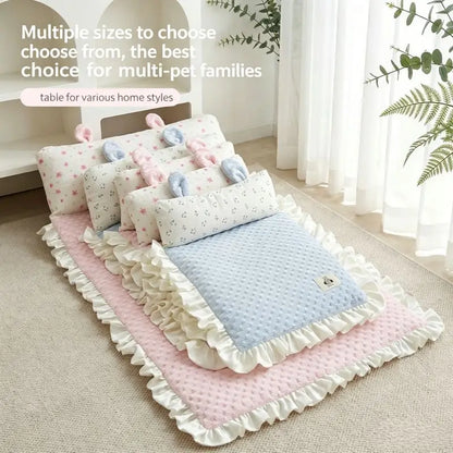 A set of soft baby bedding and pet beds arranged on a beige carpet in a bright room with white curtains and green plants. The set includes a pink ruffled mat, a light blue textured blanket, several white pillows with small prints, and pastel plush toys.