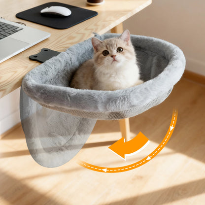 Cat sitting in a gray pet hammock on a wooden desk with a laptop and mouse.
