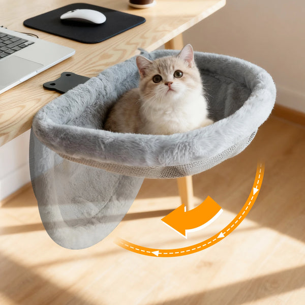 Cat sitting in a gray pet hammock on a wooden desk with a laptop and mouse.