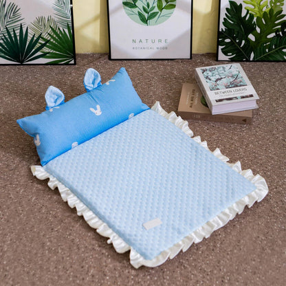 A light blue quilted baby blanket with white ruffled edges and a matching blue pillow with bunny motifs, styled on a carpeted floor with framed botanical prints and books in the background.