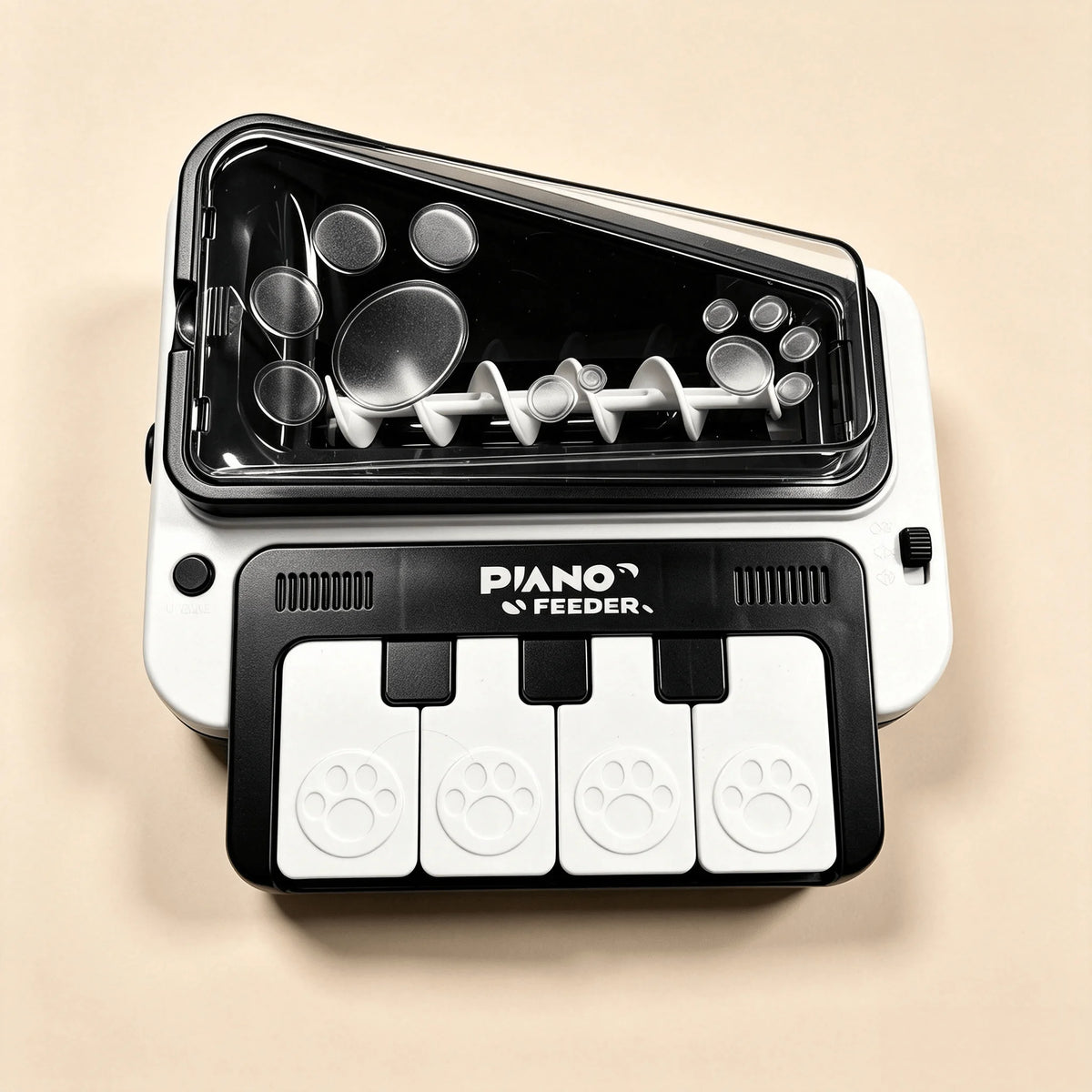 A white and black pet feeder toy shaped like a piano keyboard, with paw-print buttons and a transparent lid showing internal mixing blades, on a beige background.