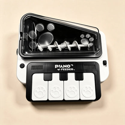 A white and black pet feeder toy shaped like a piano keyboard, with paw-print buttons and a transparent lid showing internal mixing blades, on a beige background.