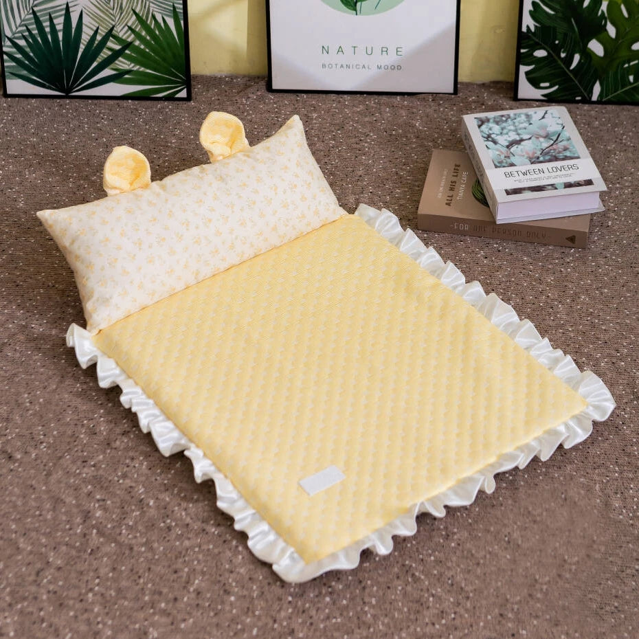A yellow quilted baby blanket with white ruffled trim and a matching pillow with yellow bunny ears, styled on a brown carpet with books and framed prints in the background.