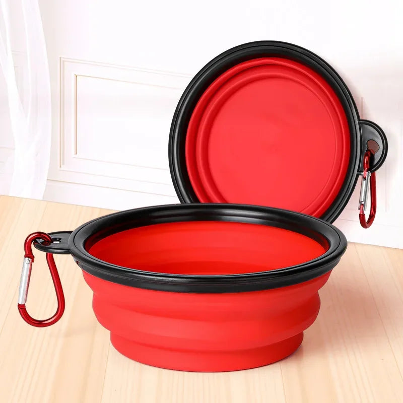 Red and black collapsible pet bowl with carabiner on a light wooden surface.