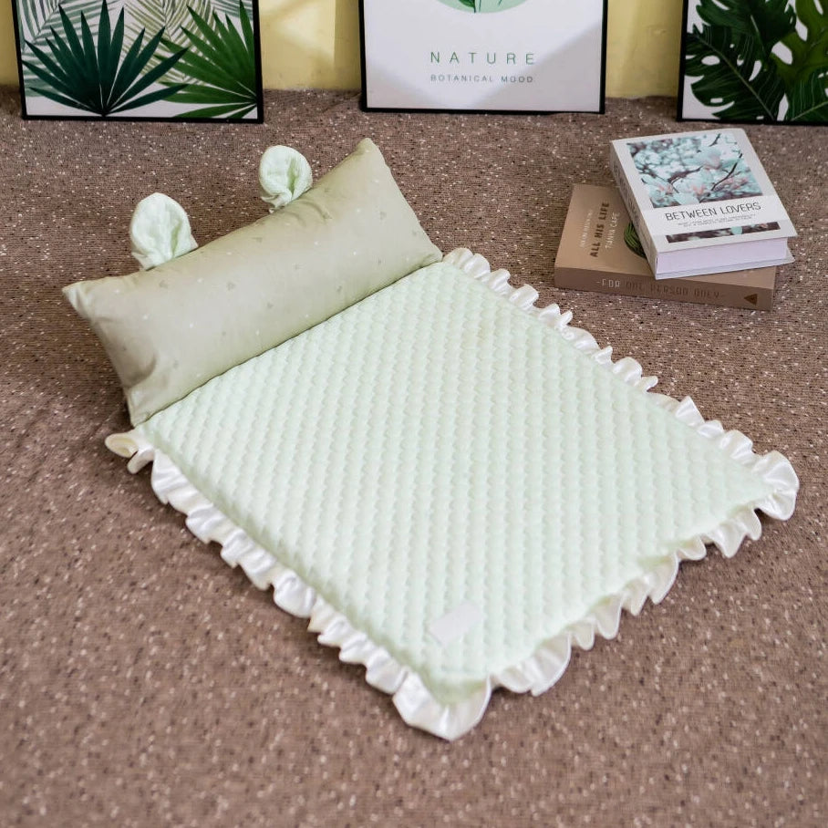 A light green quilted baby blanket with white ruffled edges and a matching pillow, styled on a brown carpet with decorative books and framed prints in the background.