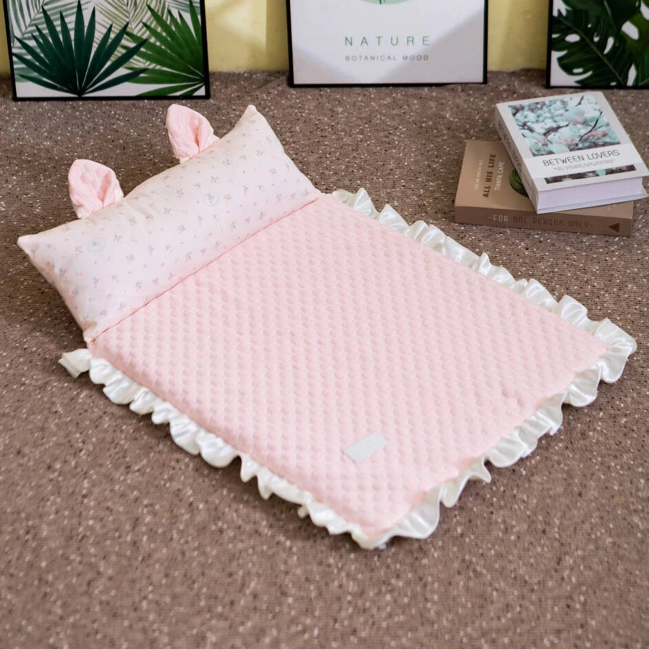 A pink quilted baby blanket with white ruffled trim and matching pink bunny-ear pillows, styled on a brown carpet with decorative books and framed prints in the background.