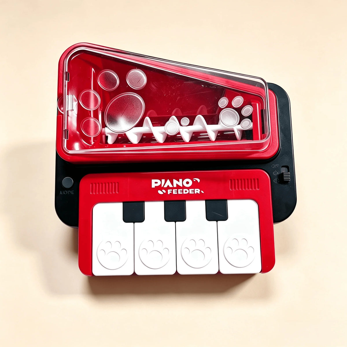 A red and black pet toy feeder shaped like a piano keyboard, with a transparent lid showing white plastic inner pieces and four white paw-print keycaps on a beige background.