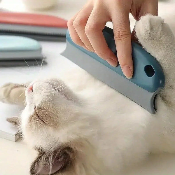A person's hand using a blue and gray pet grooming brush on a fluffy white cat lying down on a light surface.