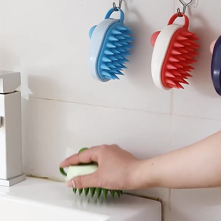 Person using a green scrubber to clean a kitchen surface with blue, red, and purple scrubbers hanging on hooks in the background.